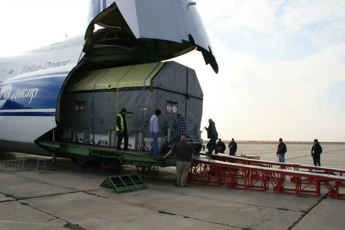 A modern cargo aircraft on an airport tarmac, nose open for loading, with ground crew and equipment visible nearby, set against a bright sky