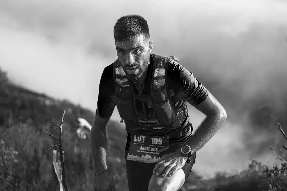 Runner's wrist wearing smartwatch on mountain trail at sunrise, terrain and sky visible in background, watch face displaying training metrics clearly visible