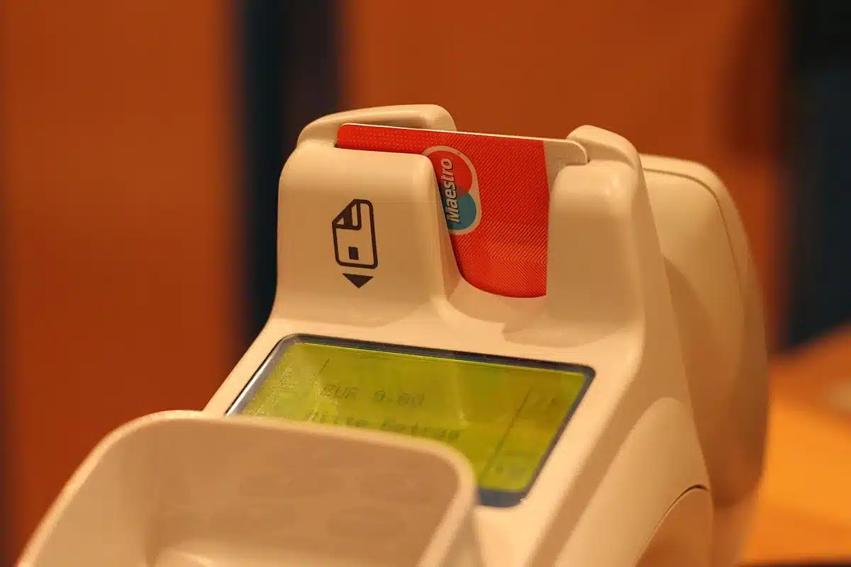A terminal point-of-sale device on a retail counter accepting a credit card, with blurred product shelves in the background, emphasizing the hidden cost of each transaction