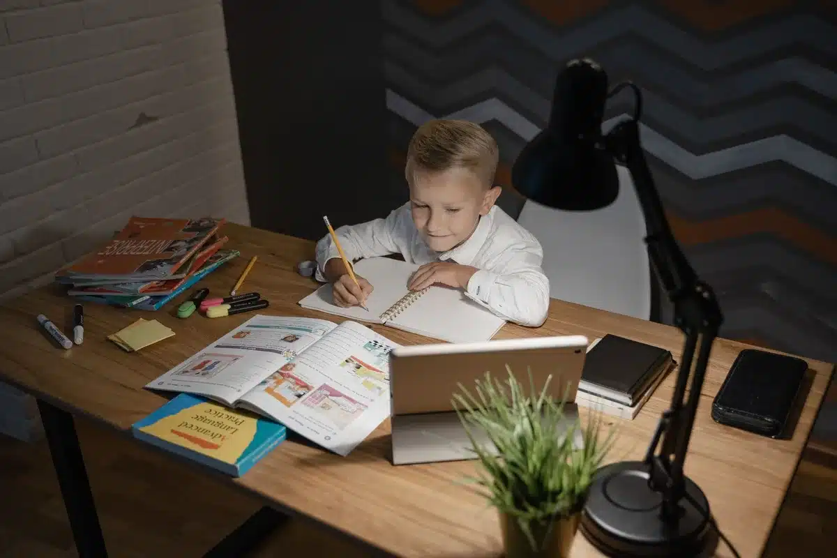Student sitting at a desk with a laptop and tablet, studying materials spread across the surface, warm natural light from a window, focused concentration