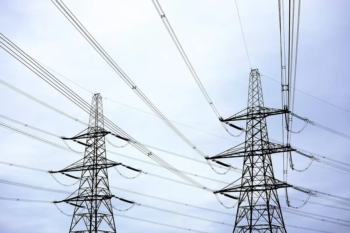 High-voltage transmission tower silhouette at dusk, damaged insulation chains visible, industrial landscape with power lines stretching across horizon, technical infrastructure damaged by vandalism, tension and disruption