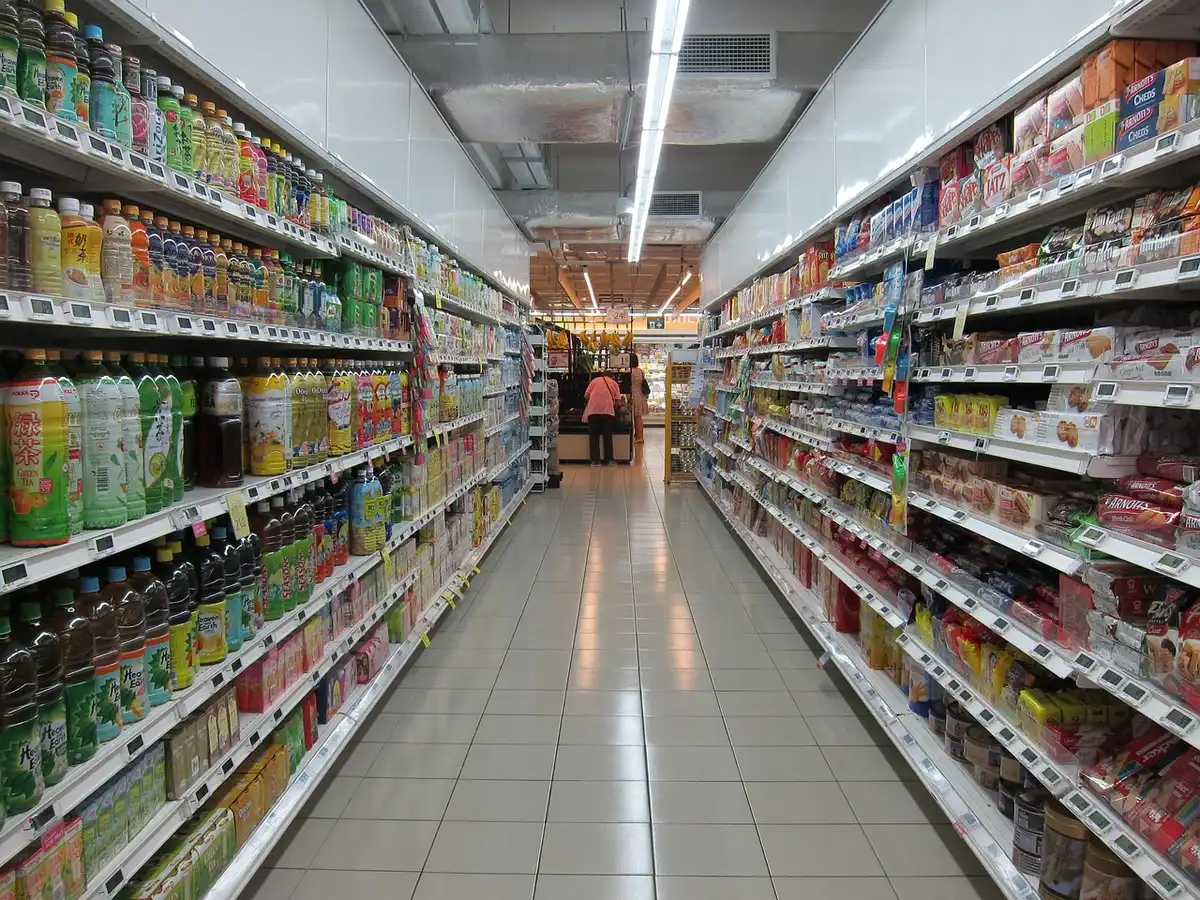 Empty supermarket aisle with stacked products and a single shopping cart, distant anonymous shopper blurred