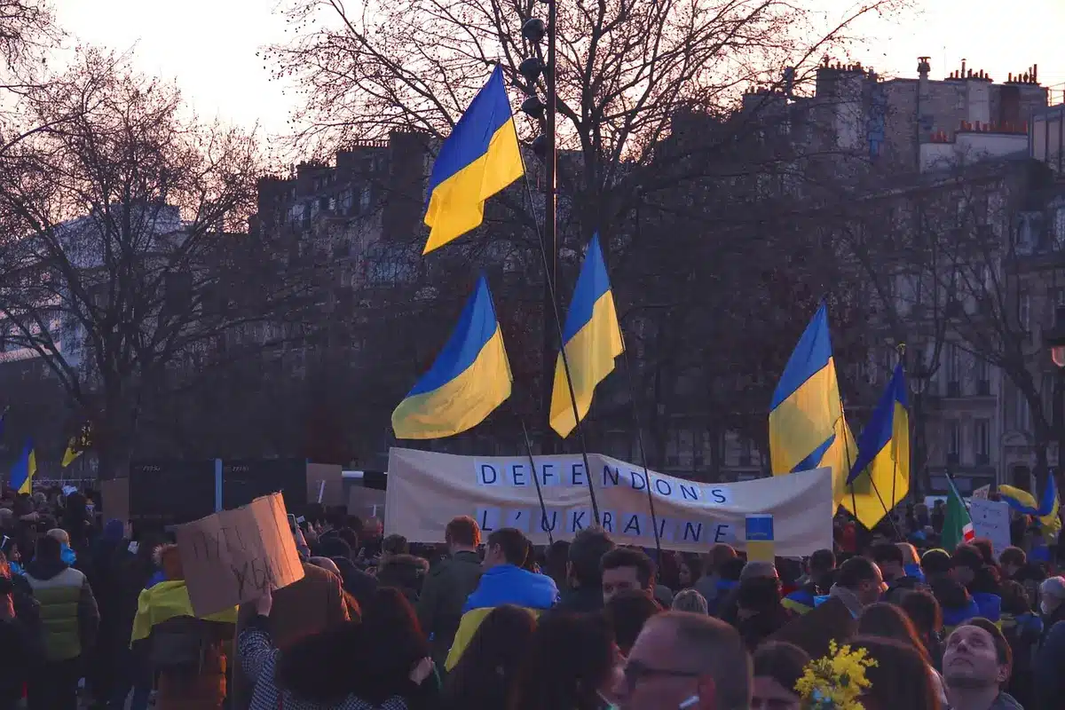 Anonymous crowd of people gathered in a public square at dusk, blurred faces, some holding smartphones aloft to record