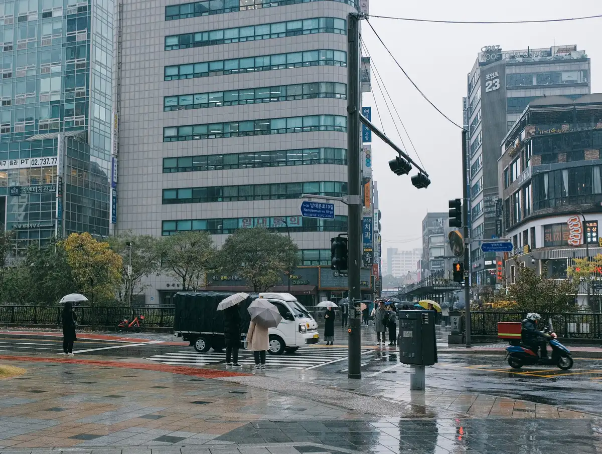 Empty street with heavy rain pooling on asphalt, blurred distant streetlights and indistinct silhouettes under umbrellas