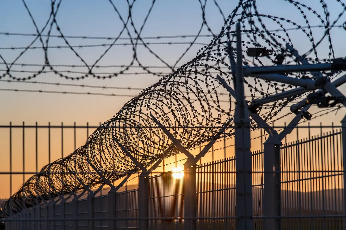 An empty high-security prison corridor with a closed cell door and a distant guard post, dim overhead lights casting long shadows