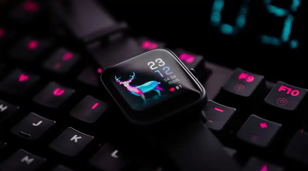 Smartwatch in premium titanium finish on a minimalist desk surface with soft morning light, showing time and date on AMOLED screen, with blurred tech accessories nearby.