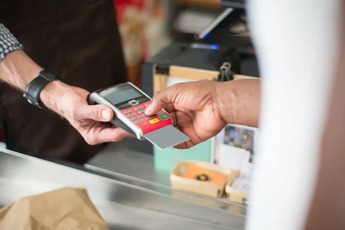 A close-up of an anonymous pair of hands tapping a generic plastic card on a compact contactless reader on a market stall table; background blurred, no logos or faces