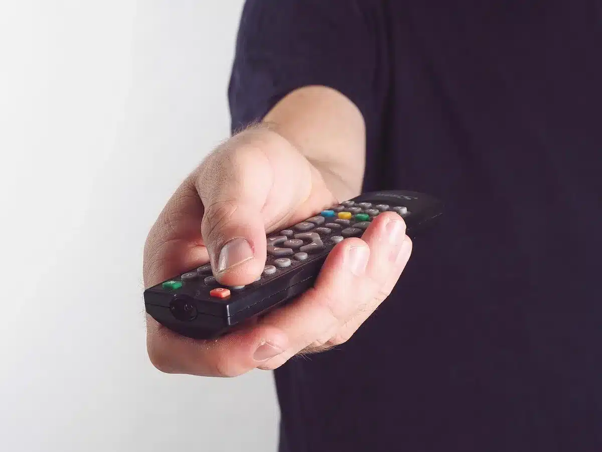 A close-up of a television screen displaying a live event with a prominent QR code overlay in the corner, with a blurred hand holding a smartphone ready to scan, warm ambient lighting