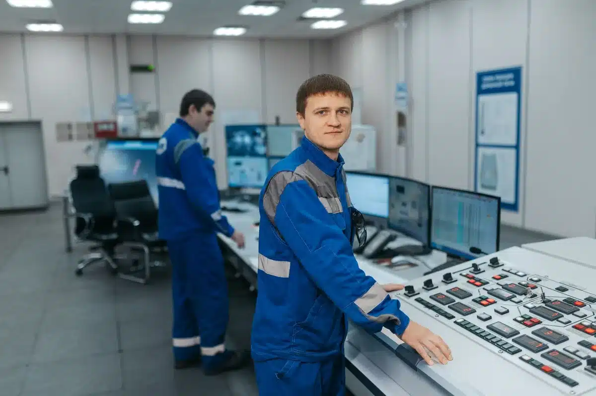 Close-up of experimental control console with anonymous hands adjusting knobs, blurred detector view through doorway, no identifying logos or text