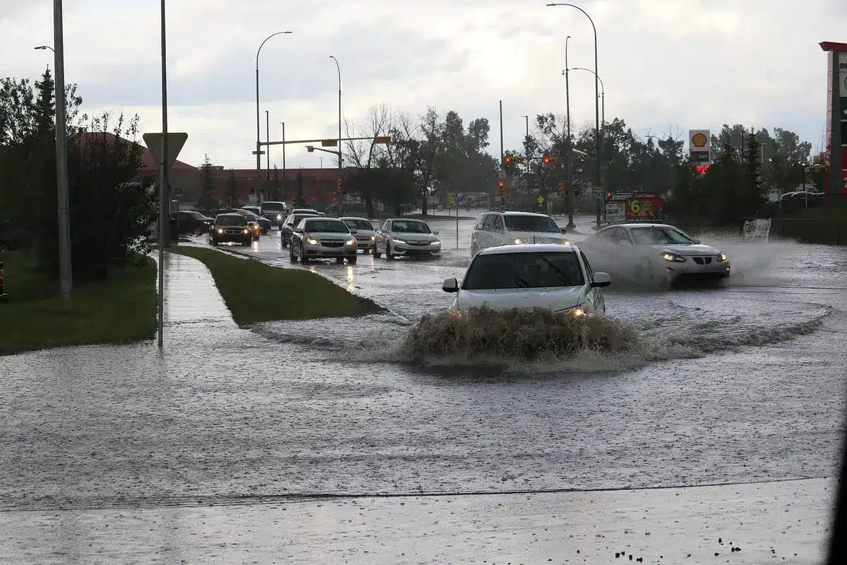 A flooded residential street with water halfway up curbs, empty of people, scattered debris near storm drains and a partially submerged motorcycle on the side