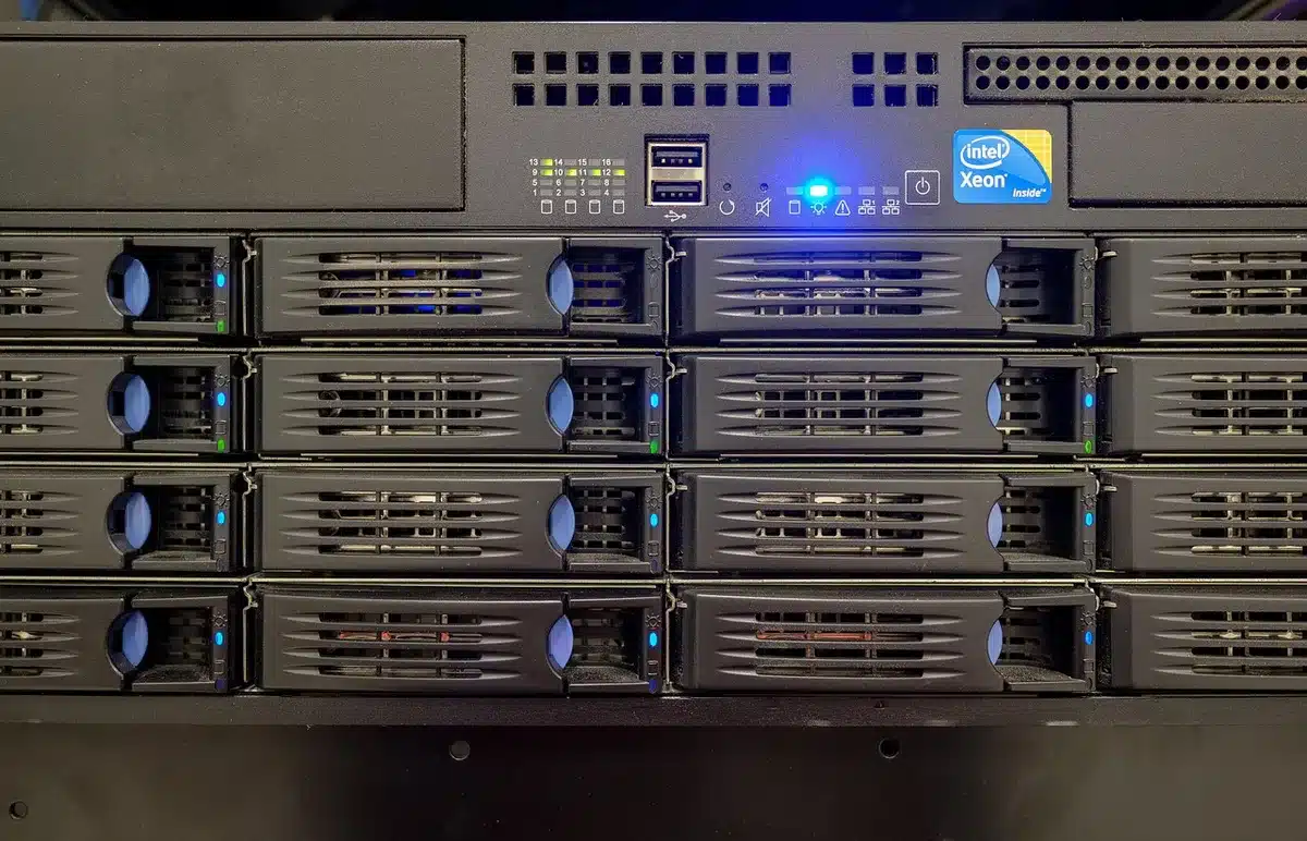 Empty server room with blinking racks and a single monitor showing a warning icon; anonymous blue-lit environment suggesting cyber risk