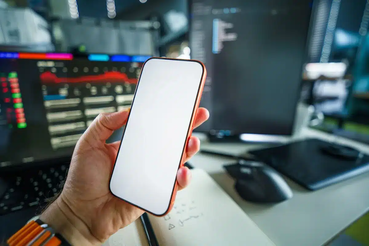 Close-up of an anonymous hand holding an unbranded smartphone displaying a generic product page with price blurred, a laptop keyboard in the background