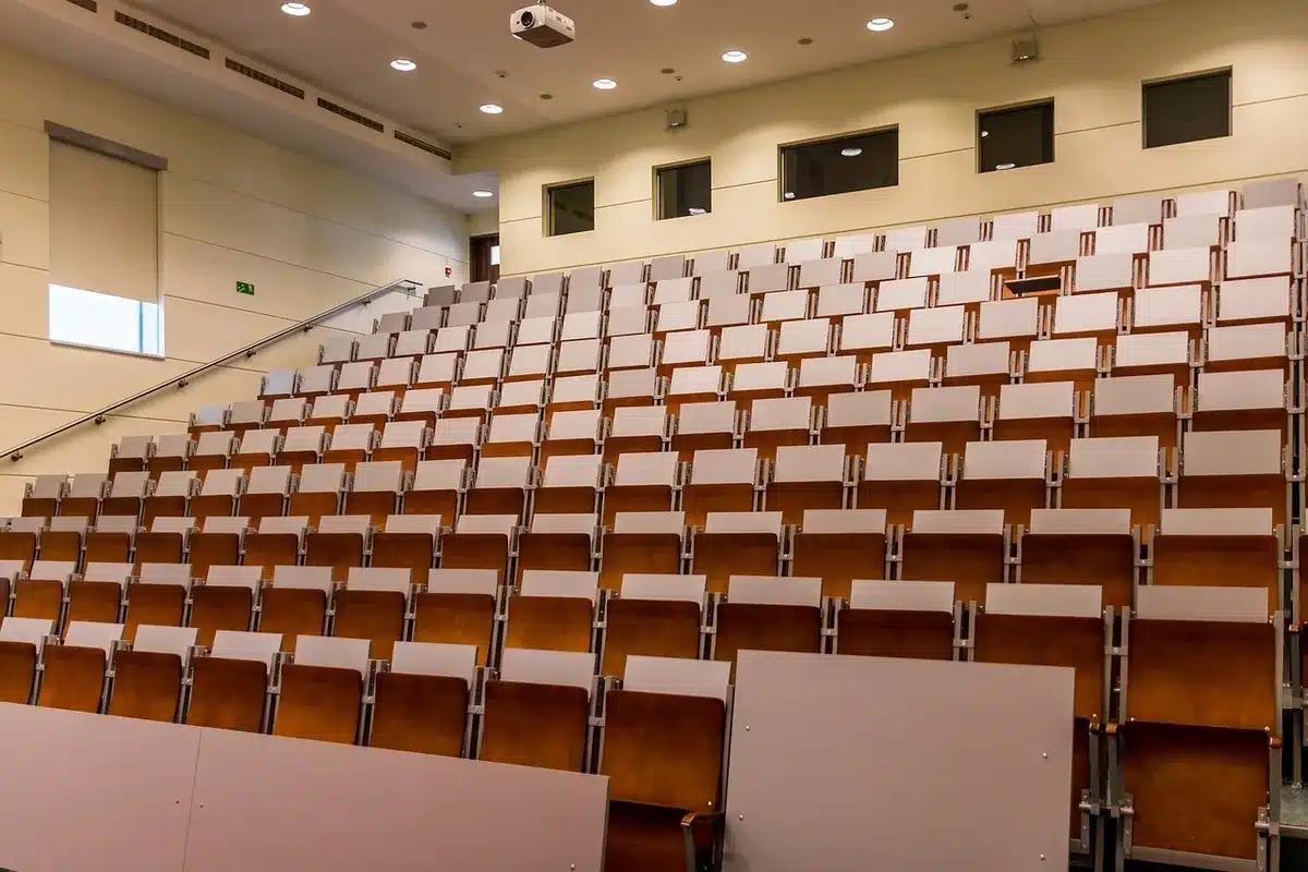 Wide shot of an empty lecture hall stage with a single microphone on a stand, scattered chairs and an academic banner in the background; no people, no logos