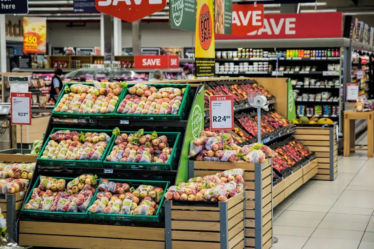 A wide produce aisle with stacked crates of anonymous apples, bananas and avocados under store lighting; no brand signs visible